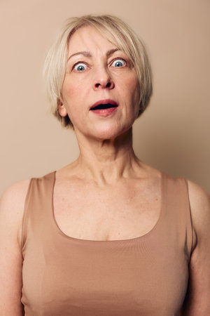 Surprised middle aged woman with short blonde hair stares wide eyed and open mouthed at camera, wearing beige sleeveless top against neutral background. Concept for shock, amazement, emotion.の写真素材