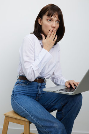 A thoughtful young woman sits on a wooden stool with a laptop, surprised expression, hand near her...