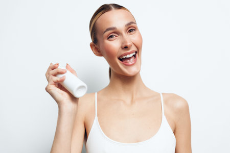 A happy young woman in a white tank top holds a white bottle while smiling brightly against a plain...