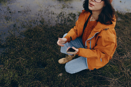 Young woman in orange jacket and glasses crouches on mossy ground near water in autumn nature....