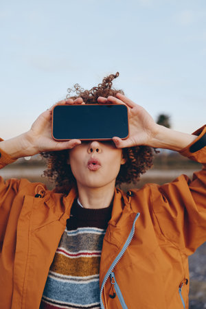 A woman with curly hair wearing a rainbow sweater and orange jacket holds a smartphone horizontally...