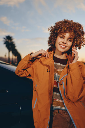 Young woman with curly hair wearing rainbow sweater and orange jacket smiles while talking on...