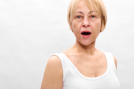 Surprised middle aged woman with short blonde hair and white tank top looks shocked with open mouth...