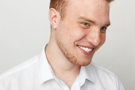 Smiling young man with red hair and freckles wearing white shirt looks down with happy expression....