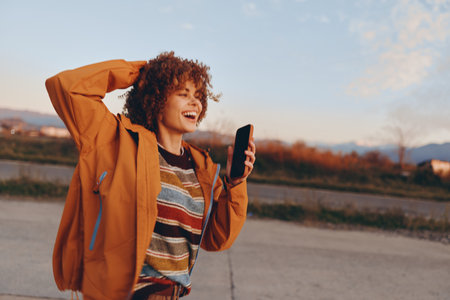 Smiling woman with curly hair wearing a rainbow sweater and orange jacket holding a smartphone...