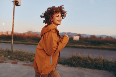 Smiling woman with curly hair wearing an orange jacket walking outdoors near a road during sunset....