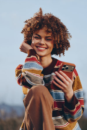 Smiling woman with curly hair wearing a rainbow sweater holding a smartphone outdoors. The woman...