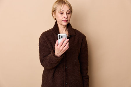 Serious middle aged woman in brown coat looks at smartphone screen on beige background. Concept of...