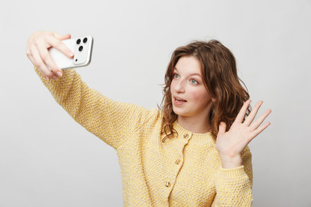 Young woman with curly hair wearing yellow sweater takes selfie with smartphone, smiling and waving...