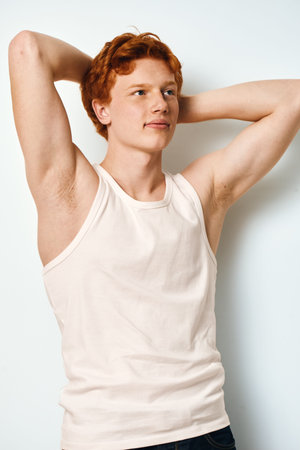 Young man with red hair and fair skin wearing white tank top posing relaxed with hands behind head...