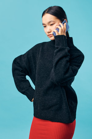 Young woman in black sweater and red skirt talking on smartphone against blue background with...