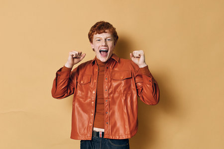 Young man with red hair smiling and celebrating with raised fists wearing brown leather jacket and...