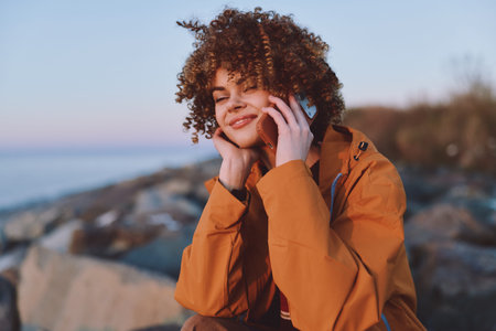 Young woman with curly hair smiles happily while talking on smartphone near rocky shore wearing...