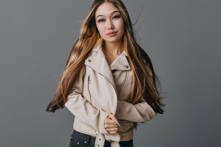 Young woman with long hair wearing beige jacket posing standing on gray background. Female model...