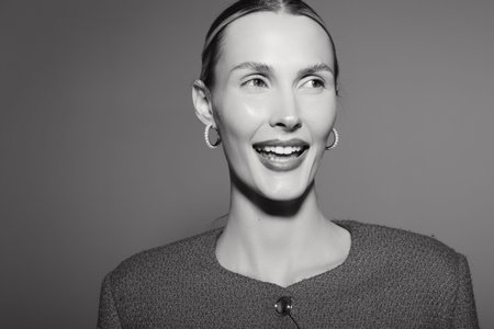 Smiling white woman with fair skin and light hair wears textured sweater and earrings against simple...