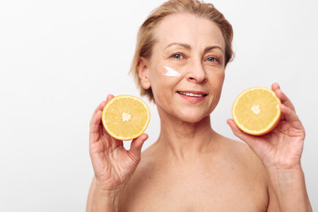 Mature woman with short blonde hair holds two halves of orange and applies facial cream on cheek....