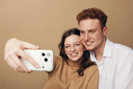 Smiling young couple takes selfie with smartphone on beige background. Happy man and woman enjoy...