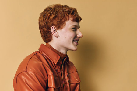 Redhead man with curly hair wearing a brown jacket smiling and looking sideways on beige background...