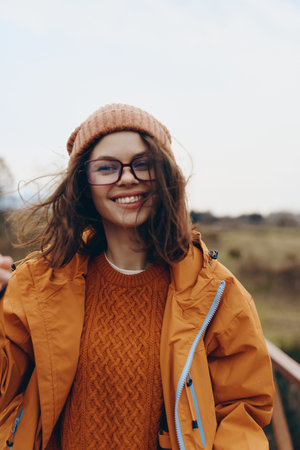 Smiling woman in orange jacket and knitted sweater enjoys outdoor lifestyle, wearing glasses and...