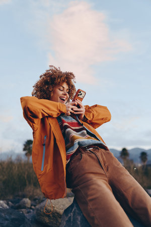 A happy woman in a colorful striped sweater and orange jacket smiles while using a smartphone...