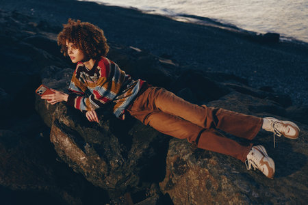 Woman with curly hair wearing a rainbow sweater and brown pants lies on rocks near water holding a...