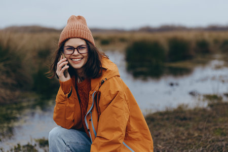 Smiling woman in orange jacket and knit hat talks on phone outdoors near a lake. Lifestyle concept showing happiness, communication, nature, autumn, and casual fashion in a natural setting.の写真素材