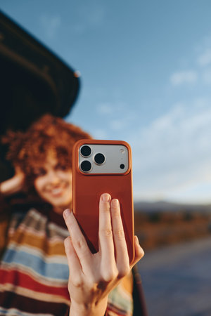 Woman with curly hair wearing a rainbow sweater takes a selfie with a smartphone outdoors near a car. Bright natural light highlights a joyful lifestyle moment with blue sky background.の写真素材