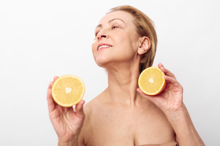 Mature woman with bare shoulders holds two halves of a grapefruit near her face, looking upwards...