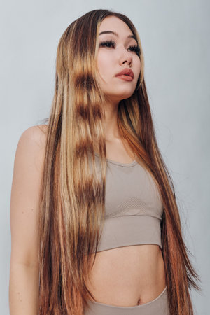 Young woman with long straight hair and natural makeup posing in beige top against light background...