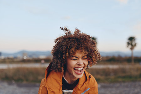 Happy woman with curly hair laughing outdoors, wearing an orange jacket, natural light, blurred background, joyful expression, casual lifestyle, sunny day, relaxed mood