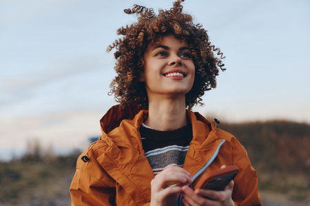A smiling woman with curly hair wearing a rainbow sweater and orange jacket holds a smartphone outdoors. The woman enjoys a casual lifestyle in a natural setting during sunset.
