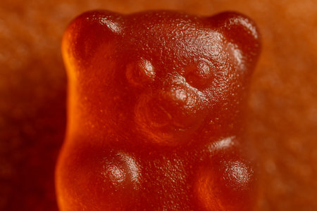 Macro photo of gummy bear surface close-up. Detailed view of shiny gelatin texture with sugar reflections and translucent candy colorの素材