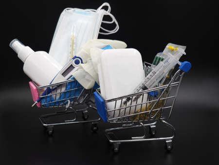 Second and third waves of coronavirus. Medical mask, disposable gloves, syringe with vaccine, antiseptic, electronic thermometer and tablets on a dark background in a shopping cart.の写真素材