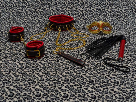 Toys for adults. Carnival mask, red and black leather handcuffs, collar, whip and nipple clamps with a gold chain on a leopard background. BDSM equipment.の写真素材