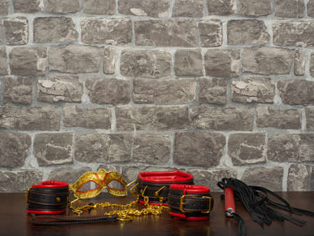 Toys for adults. A carnival mask, red-and-black leather handcuffs, a collar, a whip, and nipple clamps with a gold chain on a wooden table against a brick wall. BDSM equipment.の写真素材
