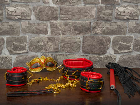 Toys for adults. A carnival mask, red-and-black leather handcuffs, a collar, a whip, and nipple clamps with a gold chain on a wooden table against a brick wall. BDSM equipment.の写真素材