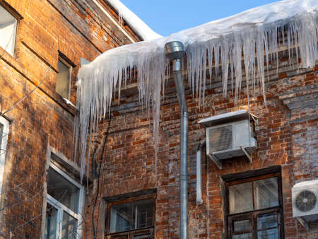 Icicles in sunlight hang on historic old building with a wall red damaged, dirty bricks. The concept of spring warming, the danger of snow falling from the roof. Facade is spoiled by air conditioning.の写真素材