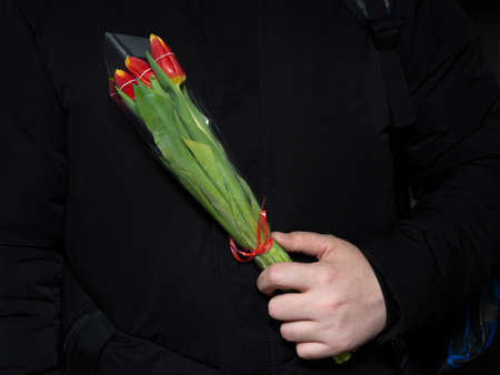 A man holds a small bouquet of tulips in a gift wrap with a red ribbon in his hand near his chest....