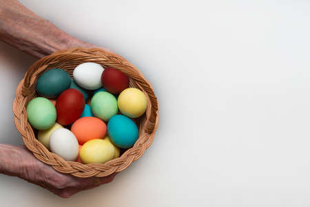 Old, elderly woman holds in her hand a multicolored, painted boiled chicken eggs in a wicker wooden,...