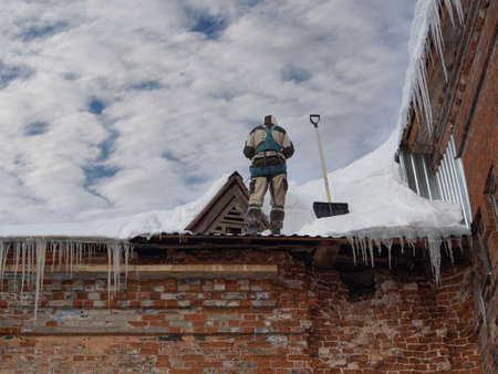 Man, industrial climber on insurance, cleans the roof of a building from snow with a shovel, a...