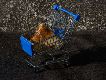 A golden, overcooked pancake folded into a triangle in a shopping cart, a trolley on a black table background on a bright sunny winter day. Traditional Russian holiday of Maslenitsa.の写真素材