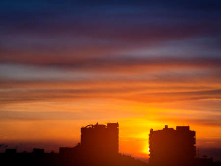 Two dark silhouettes of urban high-rise buildings against the background of an epic spring sunset in purple, red, orange and yellow shades. Glare from the sun, view at the horizon level.の写真素材