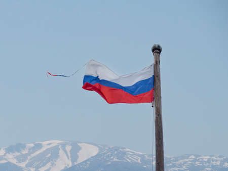 The white-blue-red flag of the Russian Federation flutters in the wind on an old wooden flagpole against the backdrop of snowy mountains. Design concept, background replacement.の写真素材