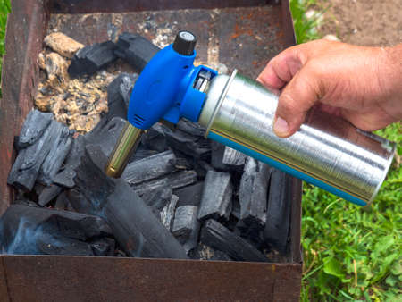 Man is holding a gas burner in his hand. Male sets fire to coals in an old rusty barbecue and makes...