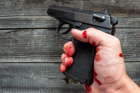 A dead man holds a police pistol in his bloody hand on a wooden background of a table or floor.