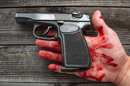 A dead man holds a police pistol in his bloody hand on a wooden background of a table or floor.の写真素材