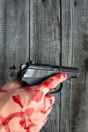 A dead man holds a police pistol in his bloody hand on a wooden background of a table or floor.の写真素材