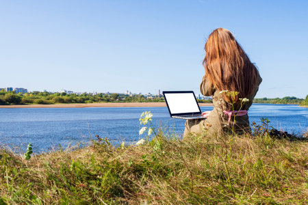 A girl with a laptop is sitting on the river bank in a park outside the city. The concept of freelance, remote work or training. An independent female businessman works in nature while traveling.の写真素材