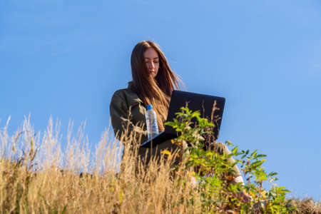 A girl with a laptop is sitting in the grass on the shore in a park outside the city. The concept of...