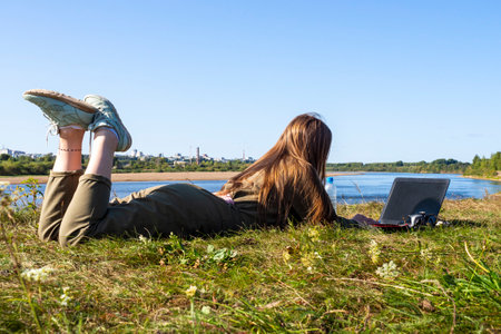 A girl with a laptop is lying on the river bank in a park outside the city. The concept of...